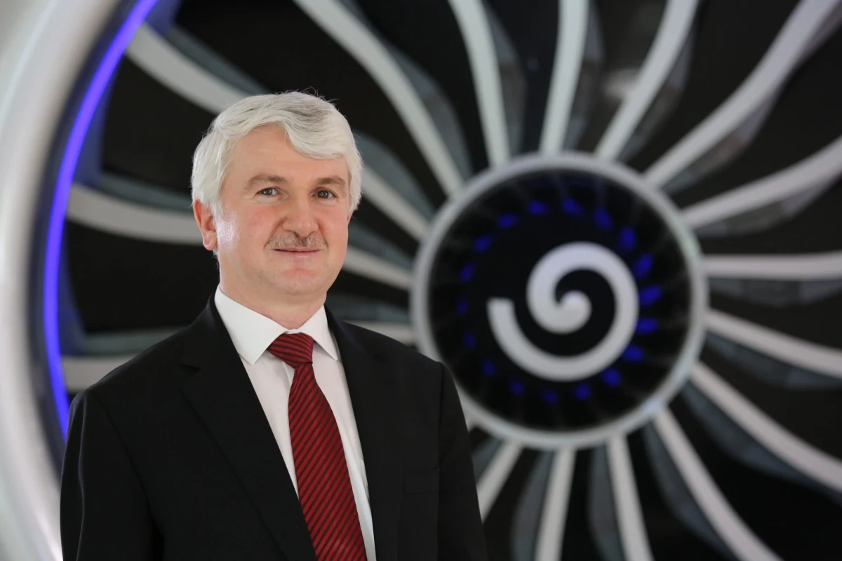 Akşit, uçak motorunu andıran büyük bir türbinin önünde takım elbiseyle poz veriyor.