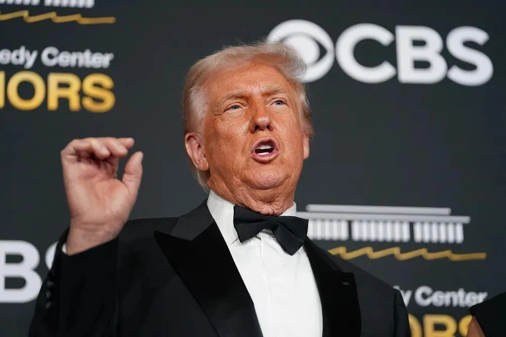 Donald Trump, siyah takım elbise ve papyonla bir etkinlikte konuşurken elini kaldırıyor.