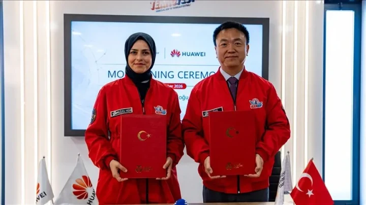 Kırmızı TEKNOFEST kıyafetleri giyen iki kişi, Huawei logosunun yer aldığı ekran önünde imzalı dosyalar tutuyor.
