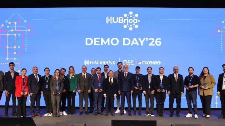 HUBrica Demo Day’26 sahnesinde etkinlik katılımcıları, arkalarında program logosunun ve Halkbank ile İTÜ Çekirdek logolarının bulunduğu mavi ekran önünde toplu halde duruyor.
